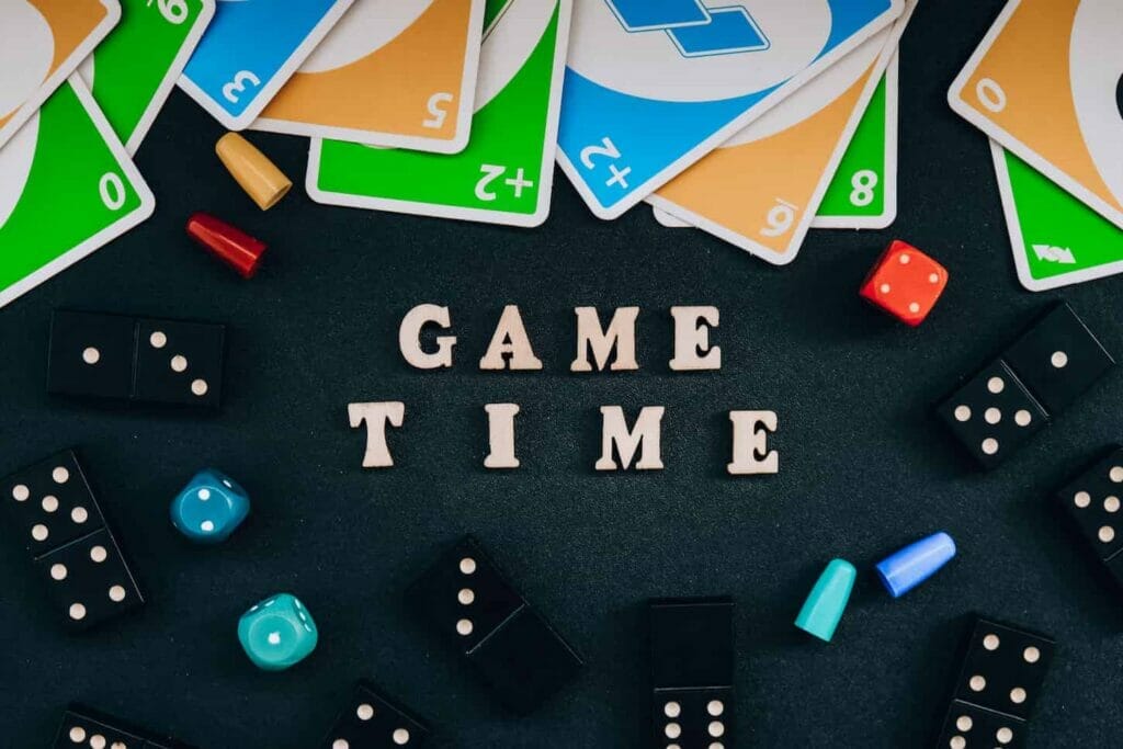 An image of Text GAME TIME spelled out in wooden letter. Surrounded by dice, dominoes uno playing cards on black.