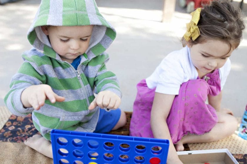 An image of two little kids playing Connect 4 game outside on a blanket.