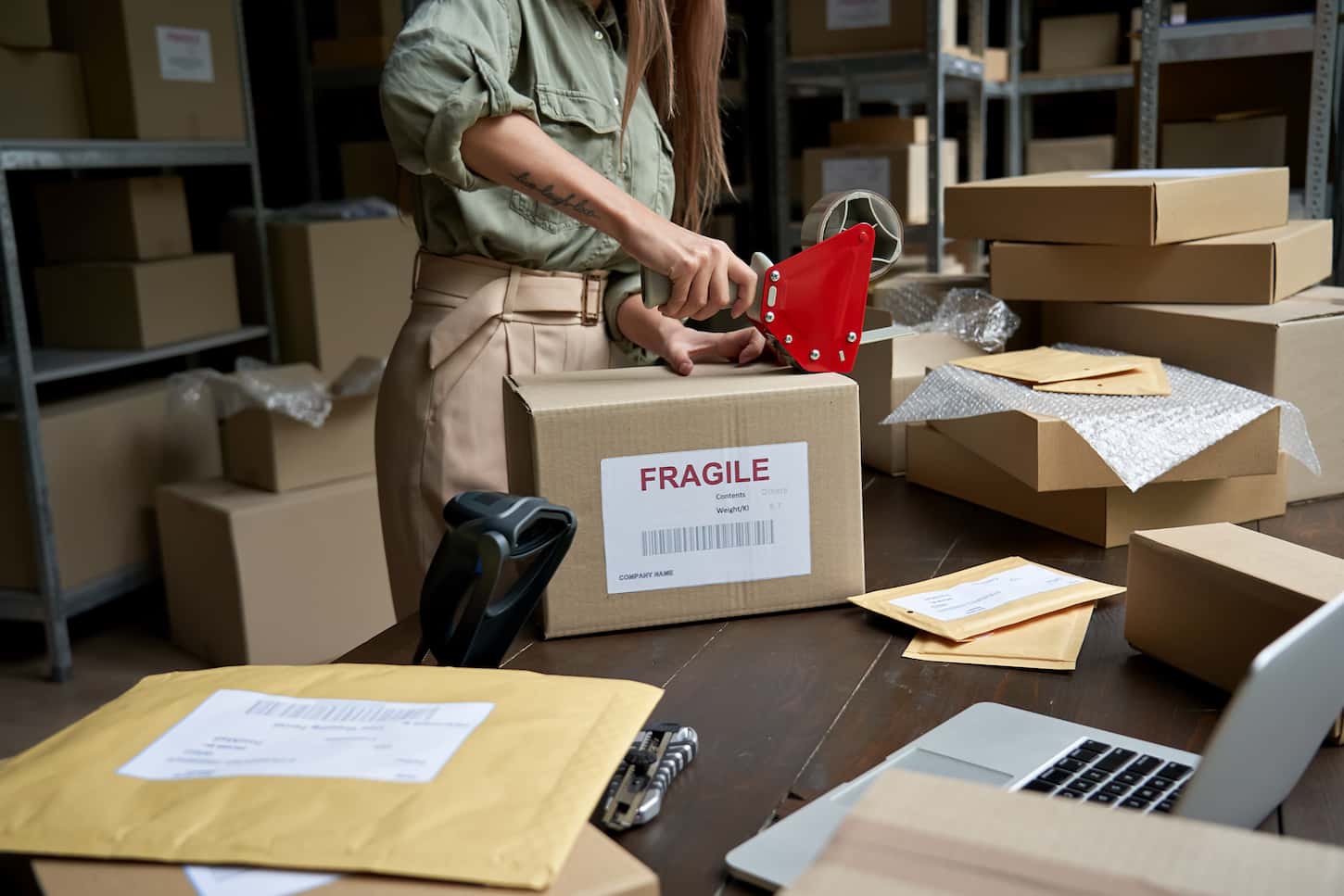 An image of a Female post mail storage worker holding a tape dispenser sealing shipping online store order courier delivery cardboard box packing e-commerce fragile dropshipping parcel in a distribution warehouse.